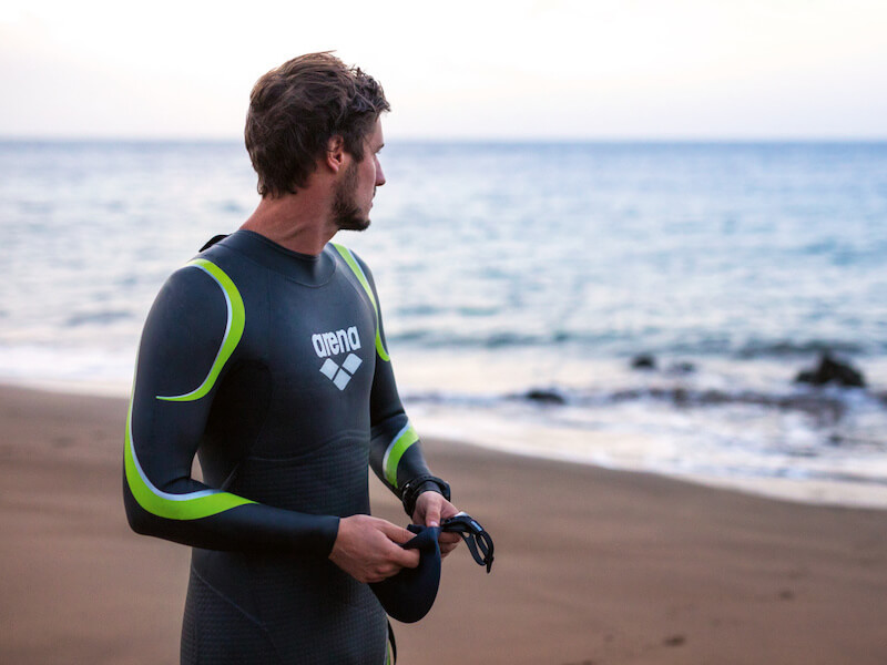 A man stands on the beach wearing an arena wetsuit